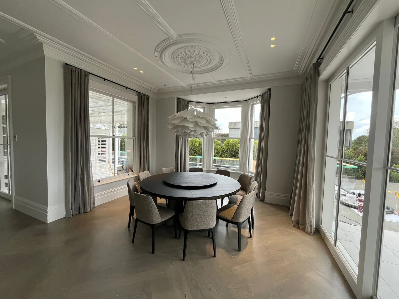 Dining room in a renovated villa painted soft grey, with panelled ceiling, ceiling rose, bay window and a round dining table